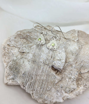 two triangular earrings on a rock with peridots in their center. there are strands of sterling silver dangling from them. 