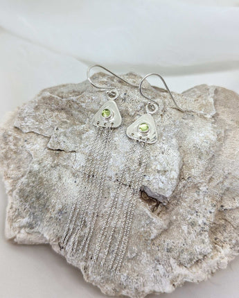 two triangular earrings on a rock with peridots in their center. there are strands of sterling silver dangling from them. 