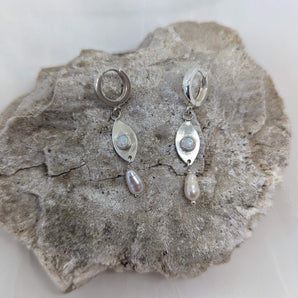 This picture shows a gorgeous sterling silver (925 silver) earrings. The earrings have dangling eye shaped pendants with an opal in their center and a pearl dangling beneath them. The earrings are presented on a white background and is shown on a rock.