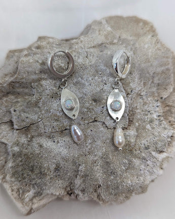 This picture shows a gorgeous sterling silver (925 silver) earrings. The earrings have dangling eye shaped pendants with an opal in their center and a pearl dangling beneath them. The earrings are presented on a white background and is shown on a rock.