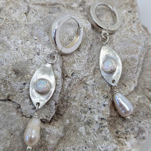 This picture shows a gorgeous sterling silver (925 silver) earrings. The earrings have dangling eye shaped pendants with an opal in their center and a pearl dangling beneath them. The earrings are presented on a rock. This picture offers a closeup image of the earrings.