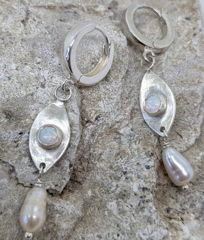This picture shows a gorgeous sterling silver (925 silver) earrings. The earrings have dangling eye shaped pendants with an opal in their center and a pearl dangling beneath them. The earrings are presented on a rock. This picture offers a closeup image of the earrings.