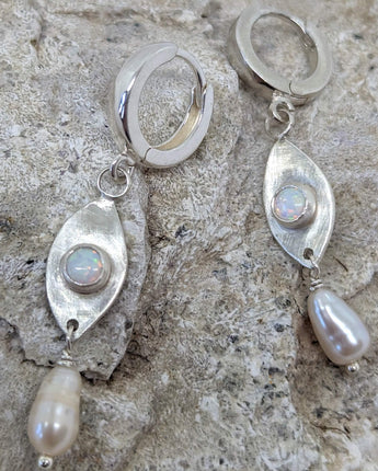 This picture shows a gorgeous sterling silver (925 silver) earrings. The earrings have dangling eye shaped pendants with an opal in their center and a pearl dangling beneath them. The earrings are presented on a rock. This picture offers a closeup image of the earrings.