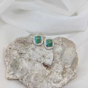 two square turquoise studs made from sterling silver presented on a rock and a white background. Farther look. 